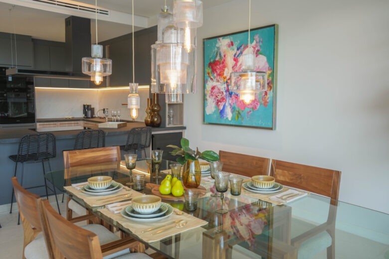 Comedor y cocina en una unidad de Macondo Playacar, con mesa de cristal y sillas de madera, vajilla artesanal, lámparas colgantes de vidrio, barra con sillas altas, gabinetes oscuros, arte colorido en la pared y detalles decorativos modernos. Espacio que refleja el estilo moderno de los departamentos en venta en Playa del Carmen dentro de este desarrollo residencial.