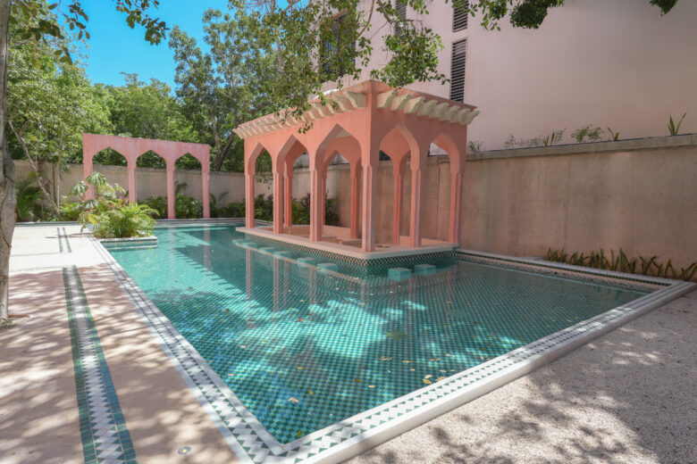 Piscina estilo riad en Pink Riad Tulum con arquitectura de inspiración marroquí en tonos rosados, detalles geométricos en mosaico verde y un pabellón central rodeado de vegetación tropical. Espacio icónico dentro de los departamentos de lujo en venta en Tulum.
